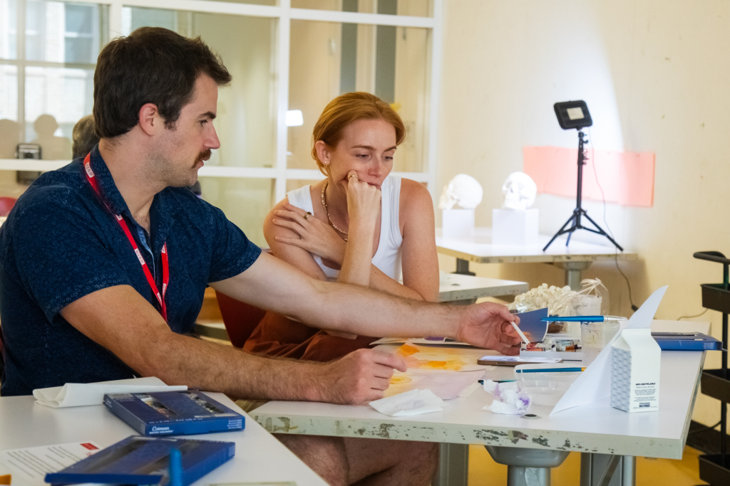 Two people sit at a table covered with art supplies and models, engaged in a creative activity. One gestures toward the materials while the other observes thoughtfully. A lamp and display items are visible in the background.