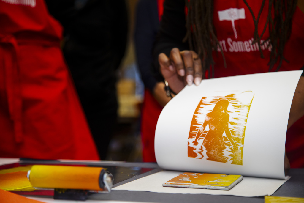 A person lifts a print of an orange and yellow silhouette of a figure from a carved block, using a printing roller, with people in red aprons visible in the background.