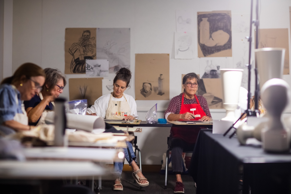 A group of people in an art classroom sketching and drawing at tables, surrounded by drawings of still life objects pinned on the wall and white sculpted objects on tables.