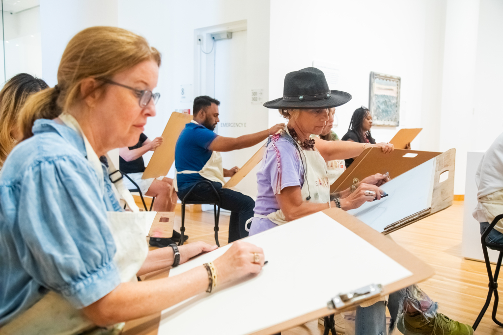 A group of adults sit in a bright art studio, each focused on sketching with pencils on large drawing boards resting on their laps. They wear aprons and are surrounded by white walls and artwork.
