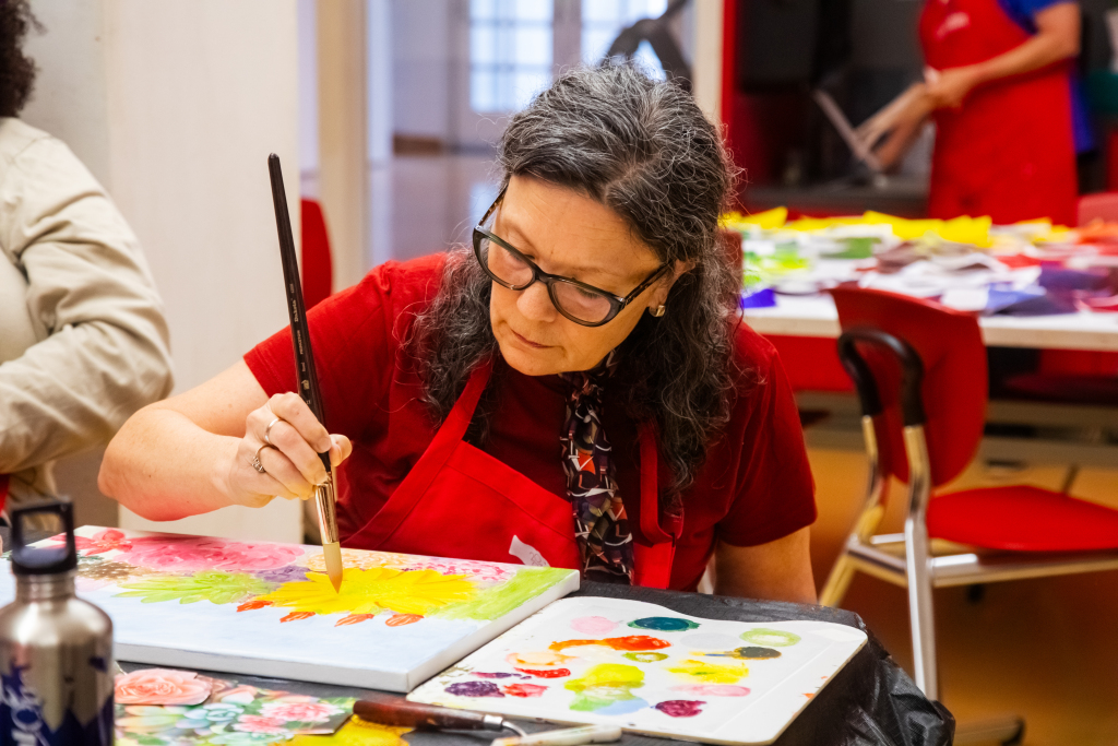 A woman wearing glasses and a red shirt paints a colorful canvas at a table, focusing intently. She is surrounded by art supplies, including palettes of paint, brushes, and a water bottle. Other people are in the background.