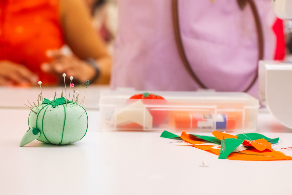 A green pincushion with pins sits on a table next to a plastic container of sewing supplies and colorful fabric scraps; people are blurred in the background.