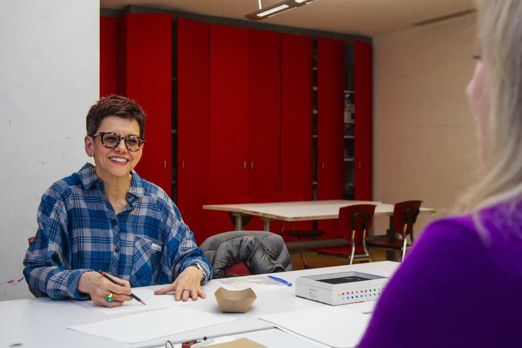 A person with short hair and glasses, wearing a blue plaid shirt, smiles while sitting at a table with papers and art supplies. Another person in purple sits across from them. Red cabinets and chairs are in the background.