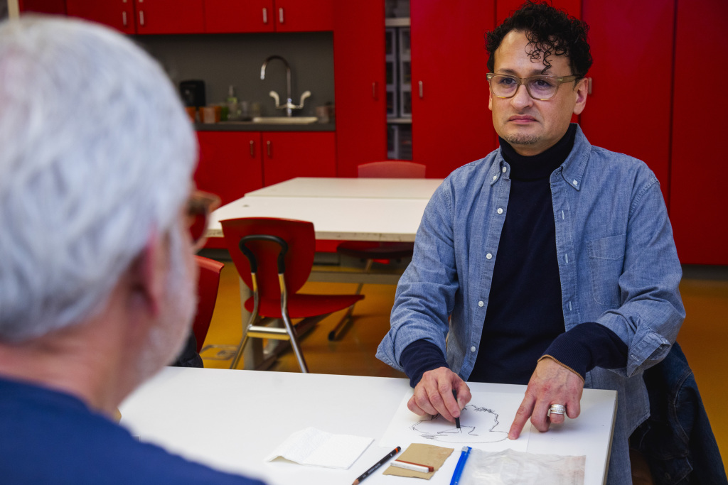A man with curly hair and glasses sits at a table, pointing to a drawing in front of him, while another person with gray hair sits across from him. Bright red cabinets and chairs are visible in the background.
