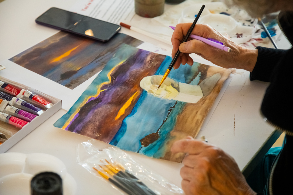 A person paints a portrait of a woman on a colorful landscape background, using watercolors. Paint tubes, brushes, a palette, and a smartphone are on the white table. The artist holds a brush over the painting.