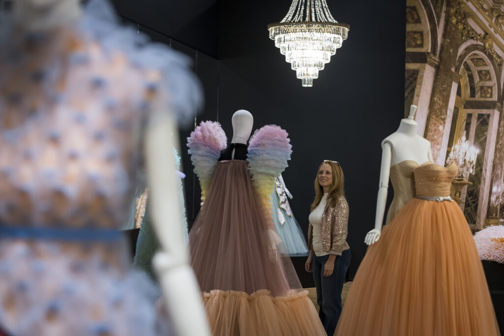 A woman smiles while viewing mannequins dressed in colorful, elegant gowns under a sparkling chandelier in a stylish exhibition space.