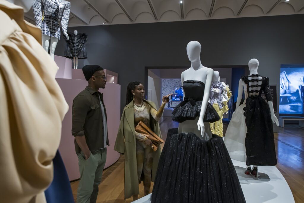 Two people observe mannequins dressed in elaborate gowns at a fashion exhibit in a museum, with various dresses displayed on platforms around them in a well-lit gallery space.