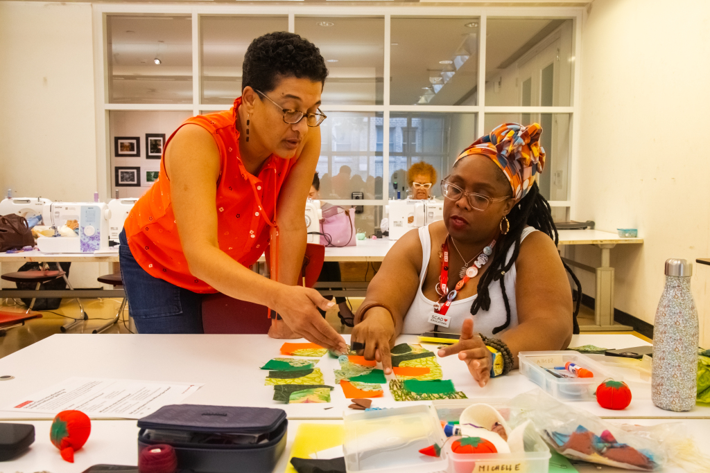 Two women sit at a table covered with colorful fabric pieces and sewing supplies. One woman stands, gesturing and explaining, while the other listens, both focused on a quilting or sewing project in a bright classroom.