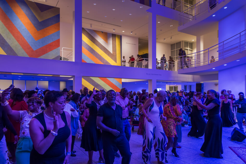 A lively crowd dances and enjoys live music in a modern, brightly lit indoor venue with colorful, geometric wall art and balconies filled with people.