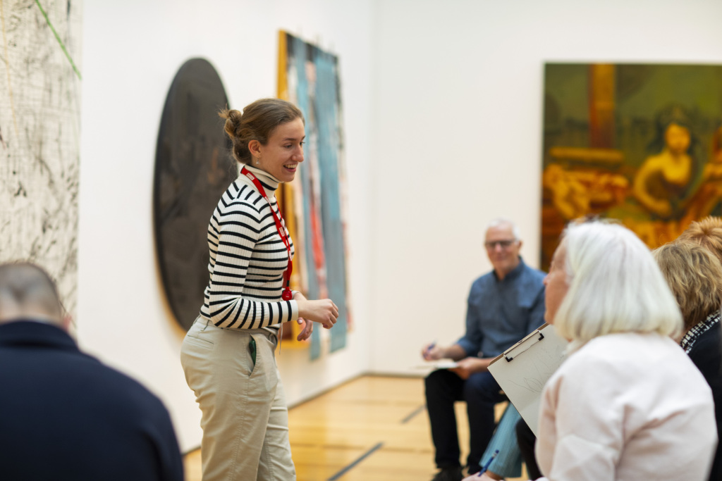 A woman in a striped shirt speaks to a seated group in an art gallery, with paintings hanging on the walls behind them. The group appears engaged, and the setting is well-lit and modern.