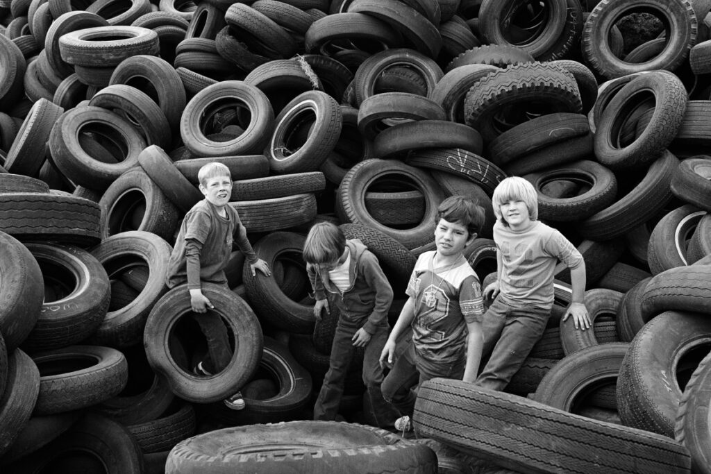 Four young boys stand and climb among a large pile of worn, stacked tires. The scene is outdoors, and the boys appear to be playing, surrounded by hundreds of overlapping tires. The image is in black and white.