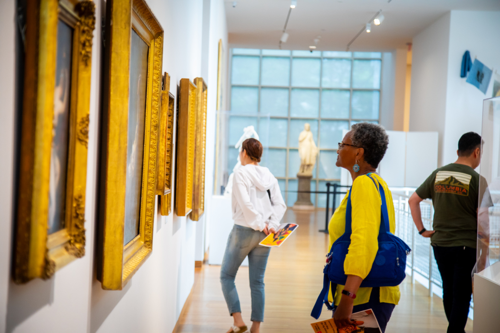 People view framed paintings in a bright art gallery. A woman in a yellow top and blue bag observes artwork closely, while others look at different pieces. A sculpture and large windows are seen in the background.