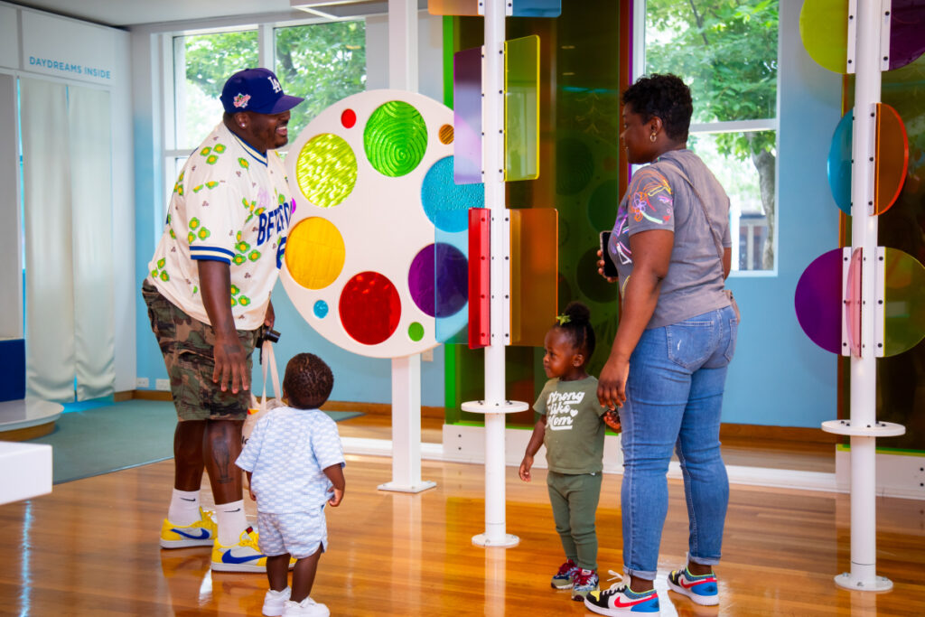 A man and woman interact with two young children in a colorful, interactive indoor play space with vibrant wall decor and large windows showing greenery outside.