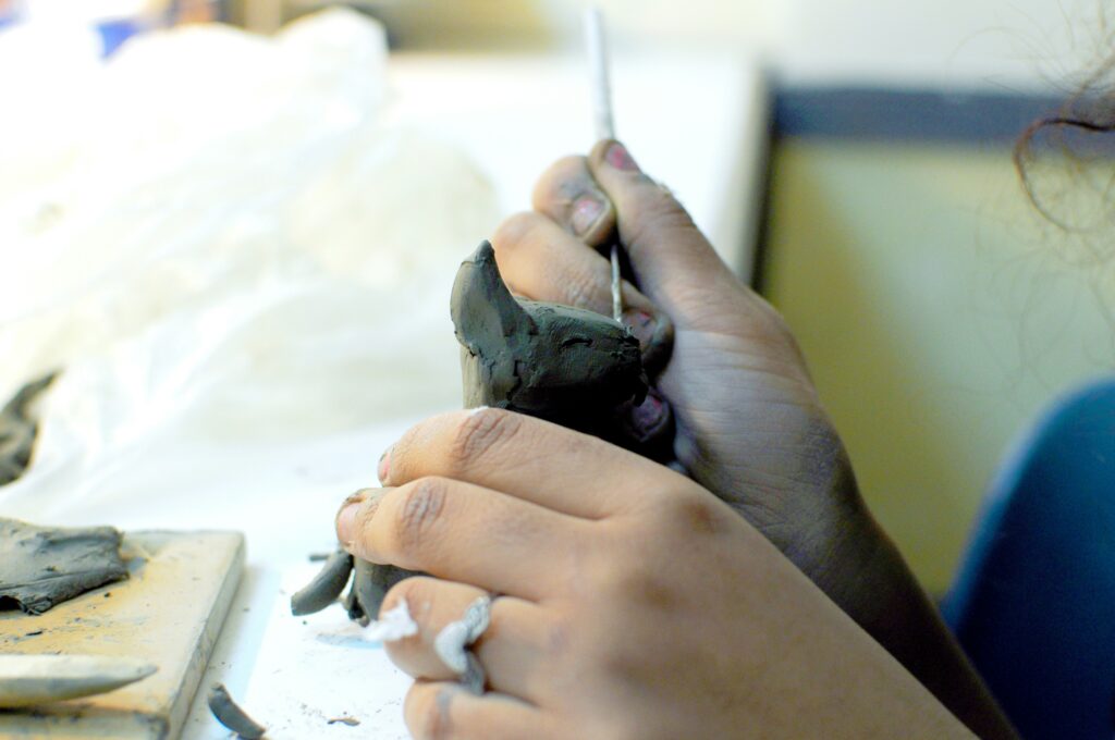 A person shapes a small clay sculpture with their hands, using a sculpting tool. The workspace is brightly lit, with pieces of clay and a cloth visible nearby.