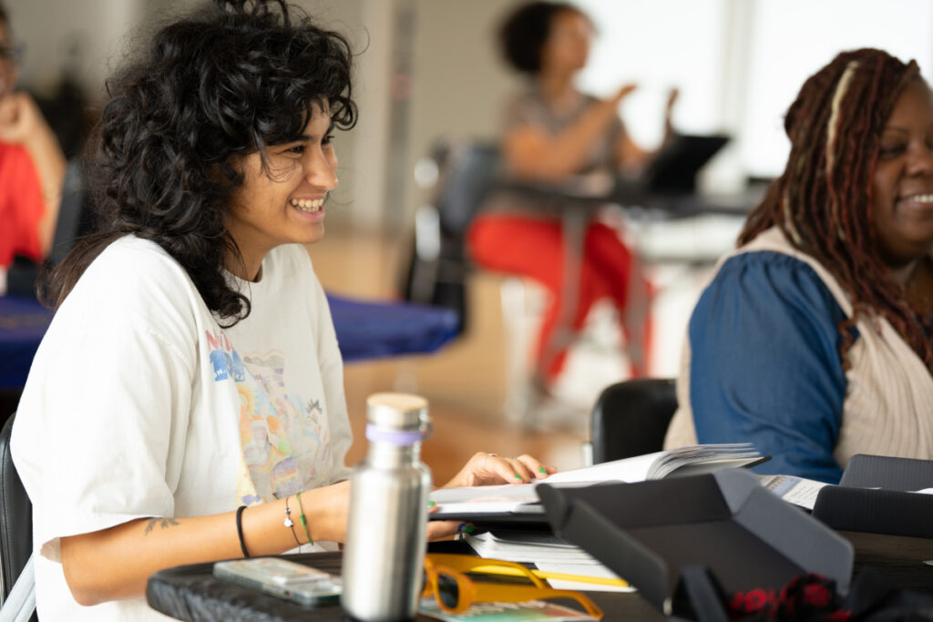 A person with curly dark hair wearing a white t-shirt smiles while sitting at a table with an open book, water bottle, and papers. Another person sits nearby, and others are blurred in the background.
