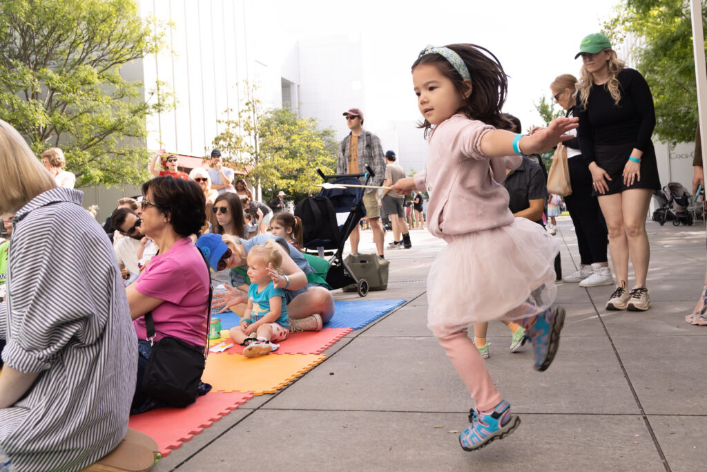 A young girl in a pink outfit twirls joyfully on a sidewalk, while adults and children sit on colorful mats and watch. People are gathered outdoors on a sunny day, surrounded by trees and modern buildings.