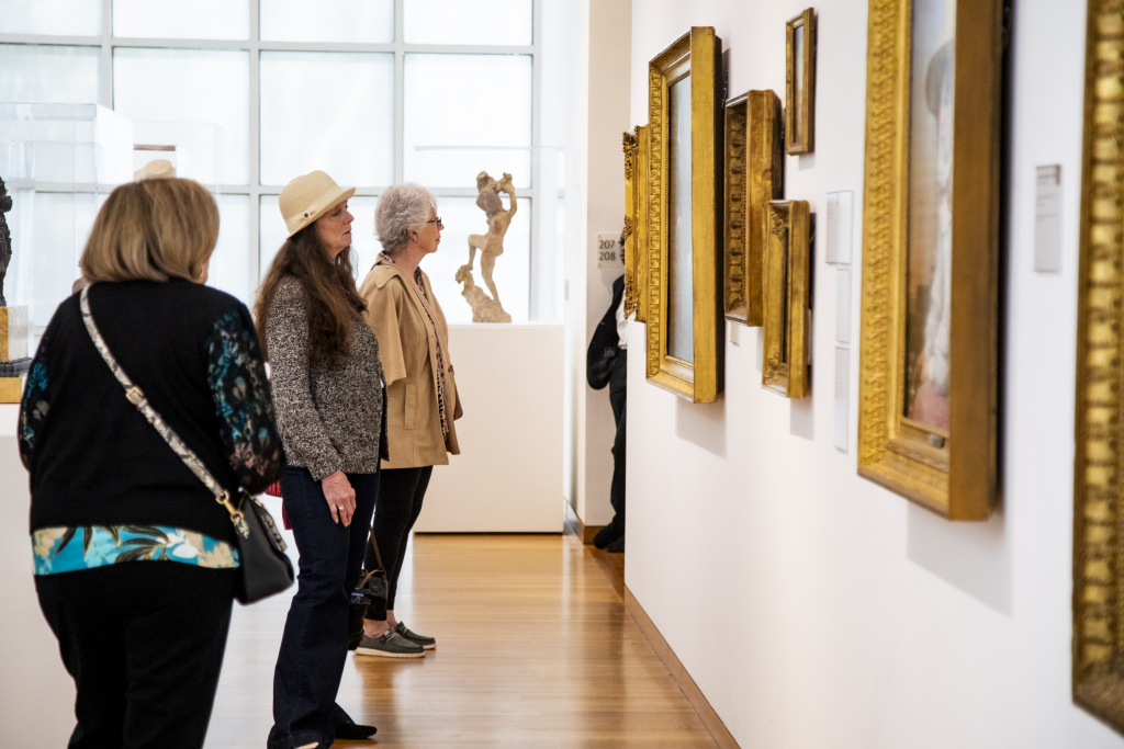 Three women stand in an art gallery, closely viewing framed paintings on a white wall. Sunlight filters through large windows in the background, and sculptures are displayed around the room.