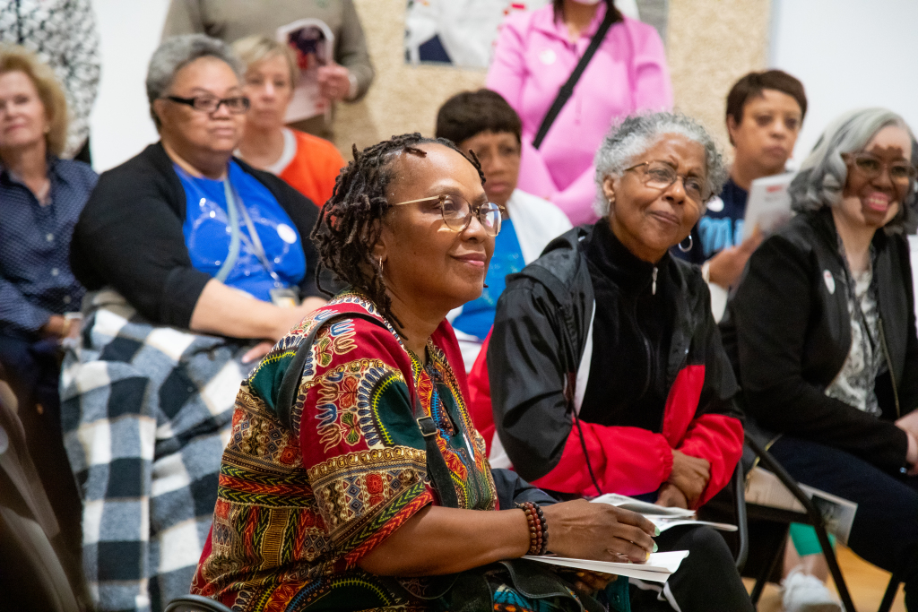 A group of women sit closely together, attentively listening to something out of frame. The focus is on two women in front, one in a colorful patterned dress and glasses, the other in a black and red jacket, both looking engaged.