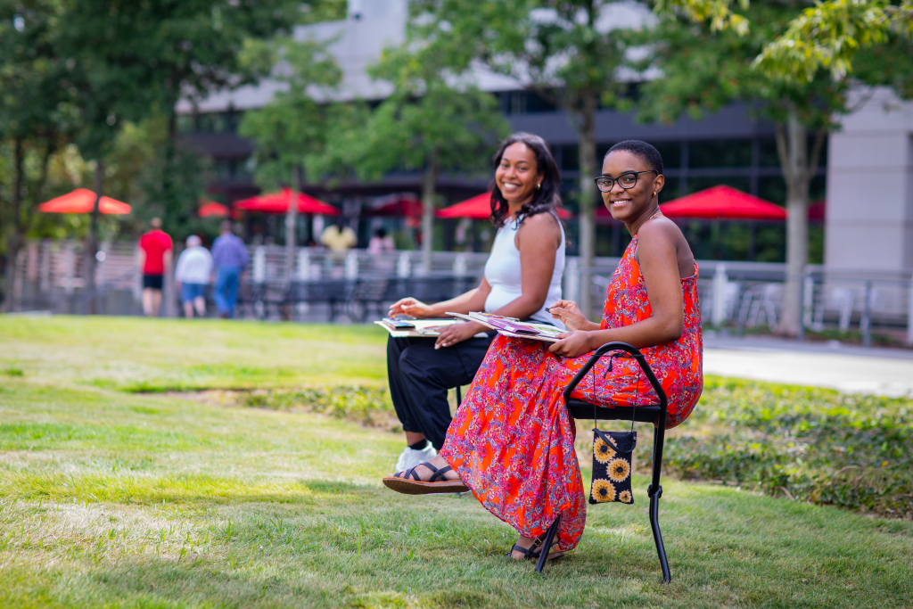 Two women sit on chairs outside on a grassy lawn, smiling and holding notebooks. One wears a white top and black pants; the other wears a bright red patterned dress. Trees and red umbrellas are visible in the background.