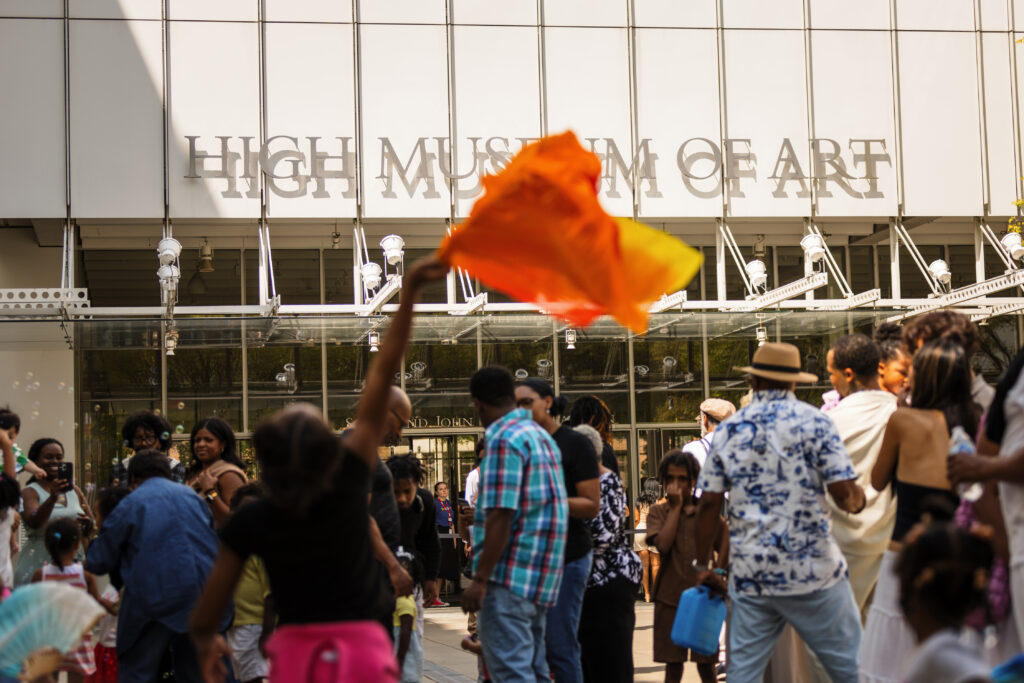 A crowd gathers outside the High Museum of Art. A person in the foreground waves an orange flag, while others socialize and walk around in the sunny outdoor area.