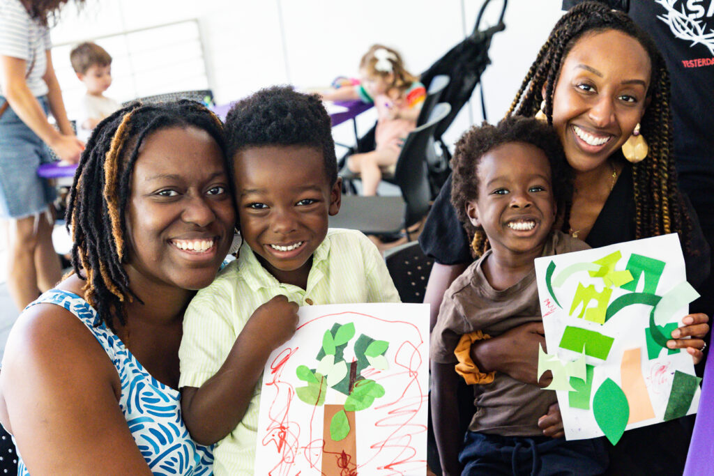 Two women and two children smiling, sitting together. The children are holding colorful, handmade art projects. Other people are visible in the background. The setting appears to be a classroom or art space.