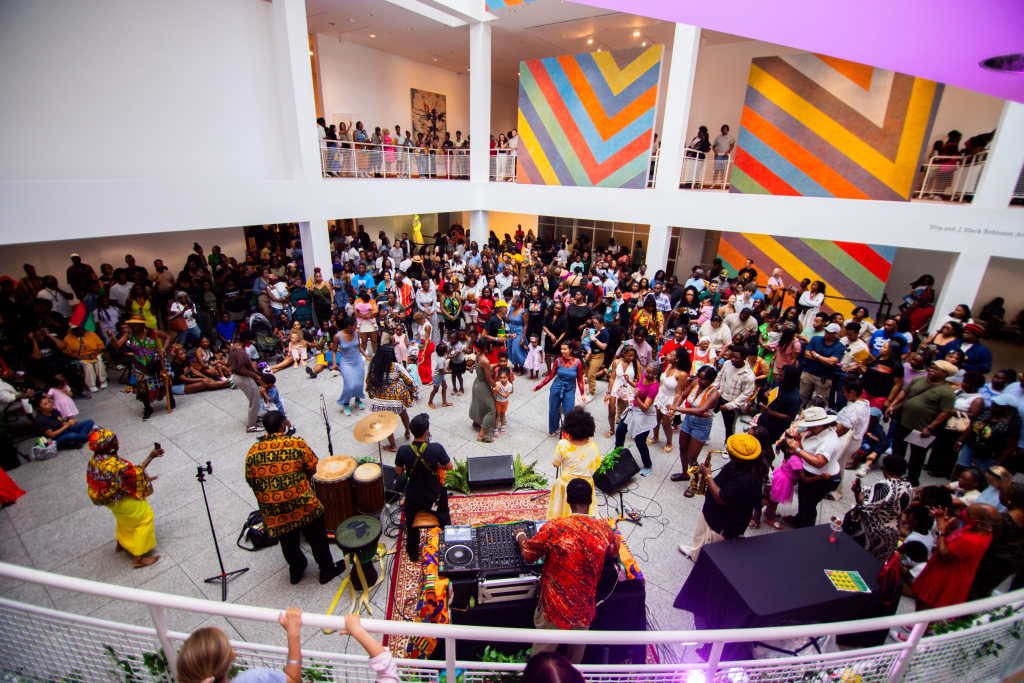 A large crowd gathers in a brightly lit atrium to watch musicians and performers, surrounded by colorful geometric wall art and two levels of spectators. Drums and DJ equipment are set up in the center.