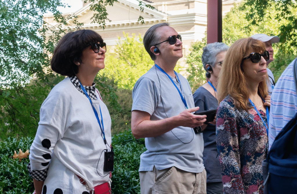 A group of adults wearing sunglasses and audio tour headsets stand outdoors, listening attentively. They have lanyards around their necks, and greenery and a building are visible in the background.