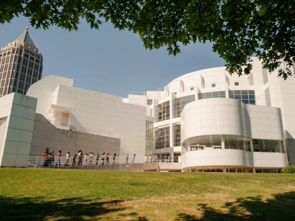 A group of people walk along a ramp outside a modern white building with curved and angular features, set against a blue sky and partially shaded by tree branches.