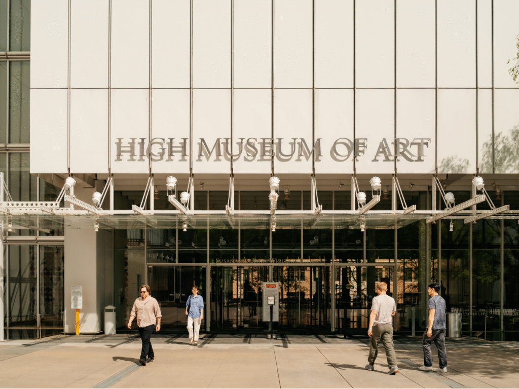Four people walk outside the entrance to the High Museum of Art, a modern building with large glass windows and the museum name displayed above the doors.