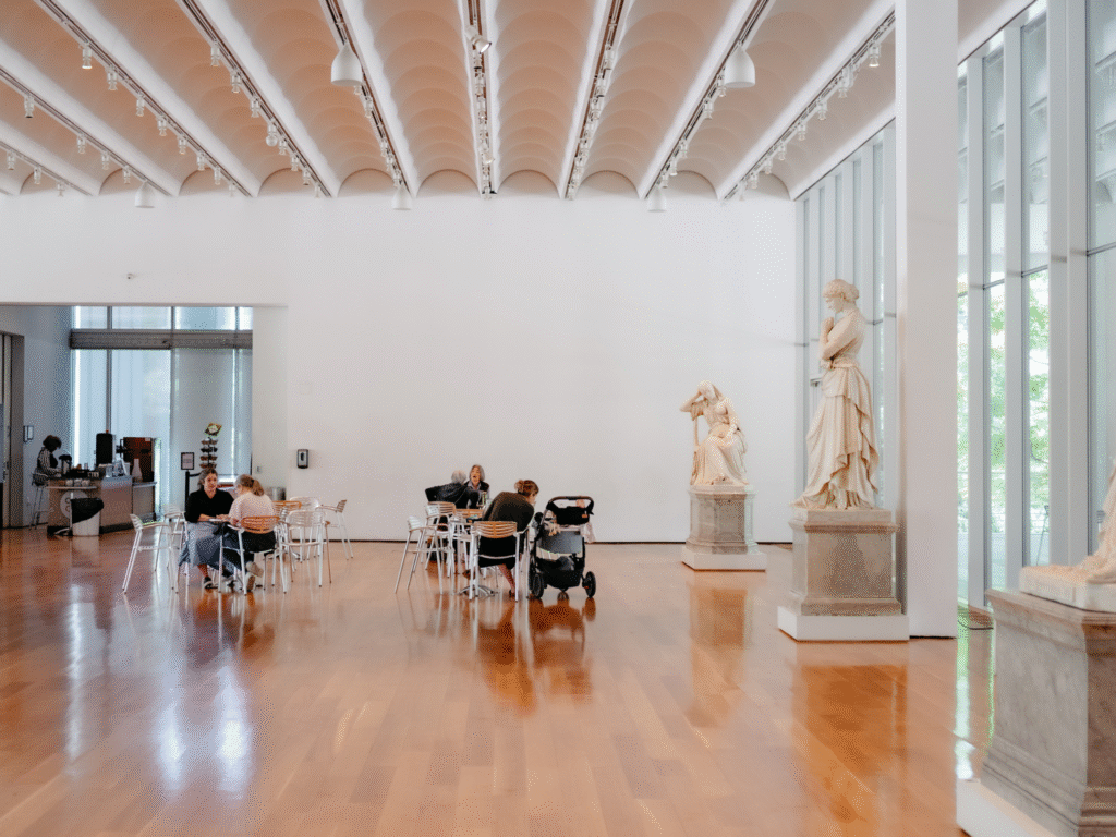 People sit at tables in a spacious, modern museum room with wooden floors, large windows, and classical white statues on display. Natural light fills the space.