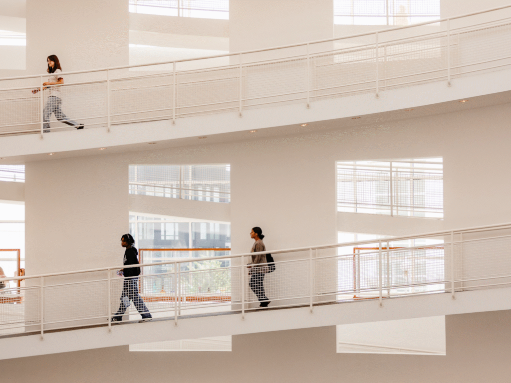 People walk along different levels of a modern building with white walls and large windows, creating a geometric, layered effect. The scene appears bright and spacious.