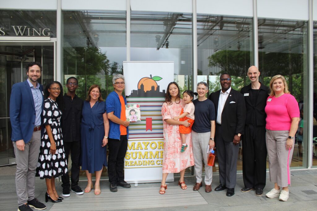 A diverse group of people stand smiling together in front of a glass building and a “Mayor’s Summer Reading Club” banner, with one woman holding a child.
