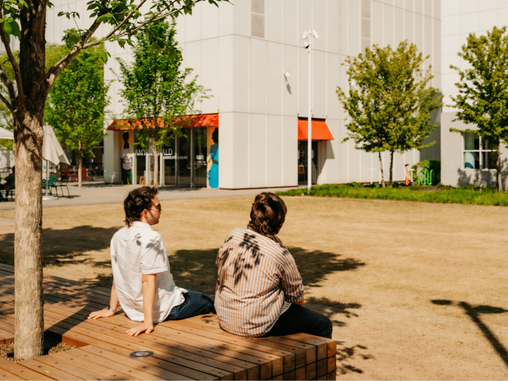 Two people sit on a wooden bench under a tree in a sunny courtyard with patchy grass. Behind them is a modern building with orange awnings and green chairs, and several small trees are scattered around.