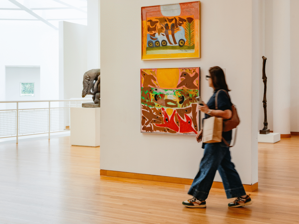 A person with a bag walks through an art gallery with wooden floors, passing colorful abstract paintings on the wall. Sculptures are visible in the background.