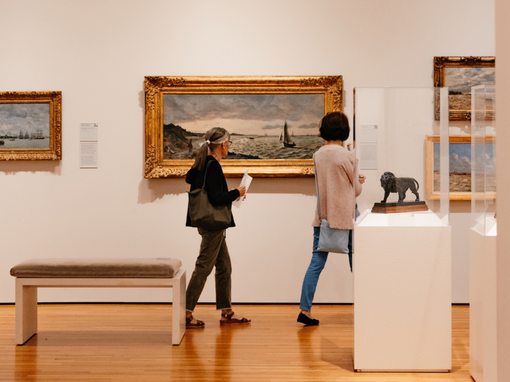 Two people view art in a museum gallery. One examines a framed painting on the wall, while the other looks at a small lion sculpture in a glass case. The room has wooden floors and a bench.