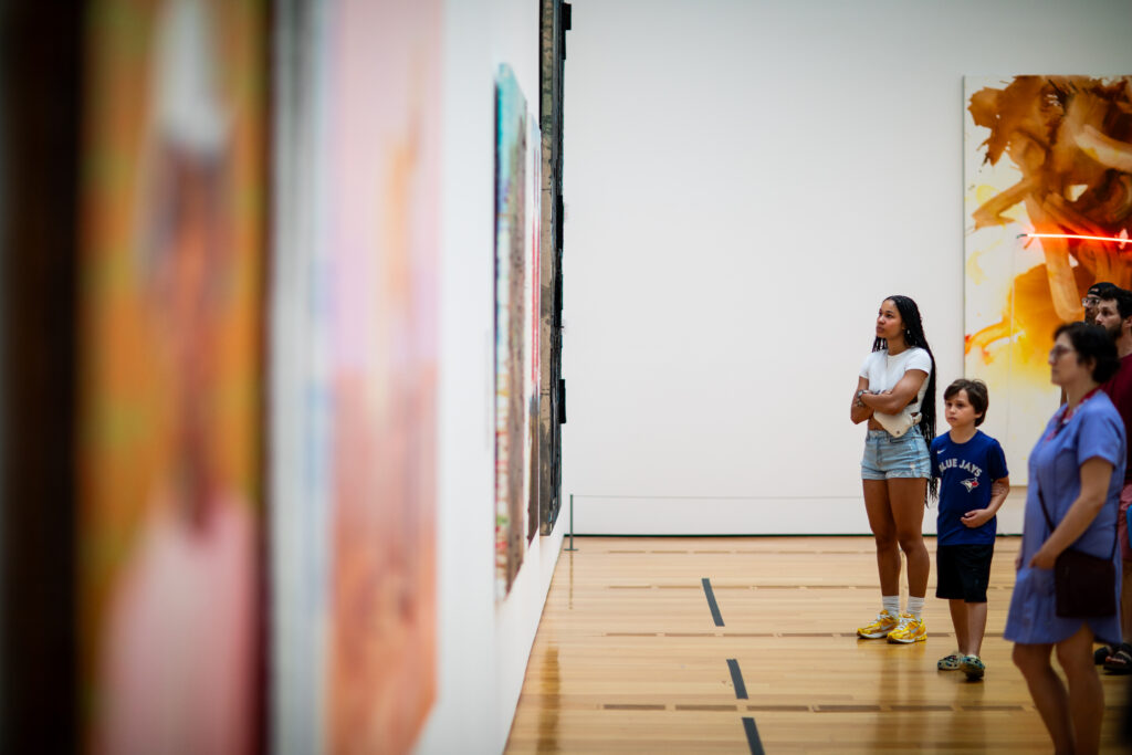 A group of people, including a woman, a boy, and two others, stand in a brightly lit art gallery, looking attentively at colorful artworks displayed on a white wall.