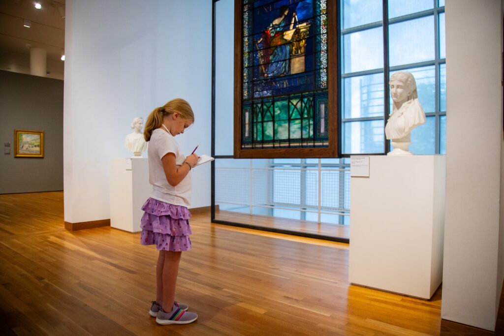 A young girl stands in an art museum, sketching on a notepad in front of a white marble bust and a tall stained glass window. The museum has wooden floors and artwork displayed on white walls.