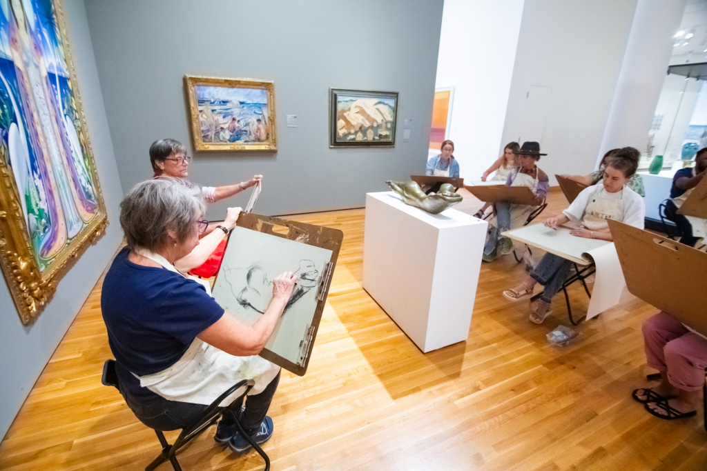 A group of adults sketching on drawing boards in an art gallery, focusing on a sculpture. Paintings hang on the walls, and the participants sit on stools around the sculpture.