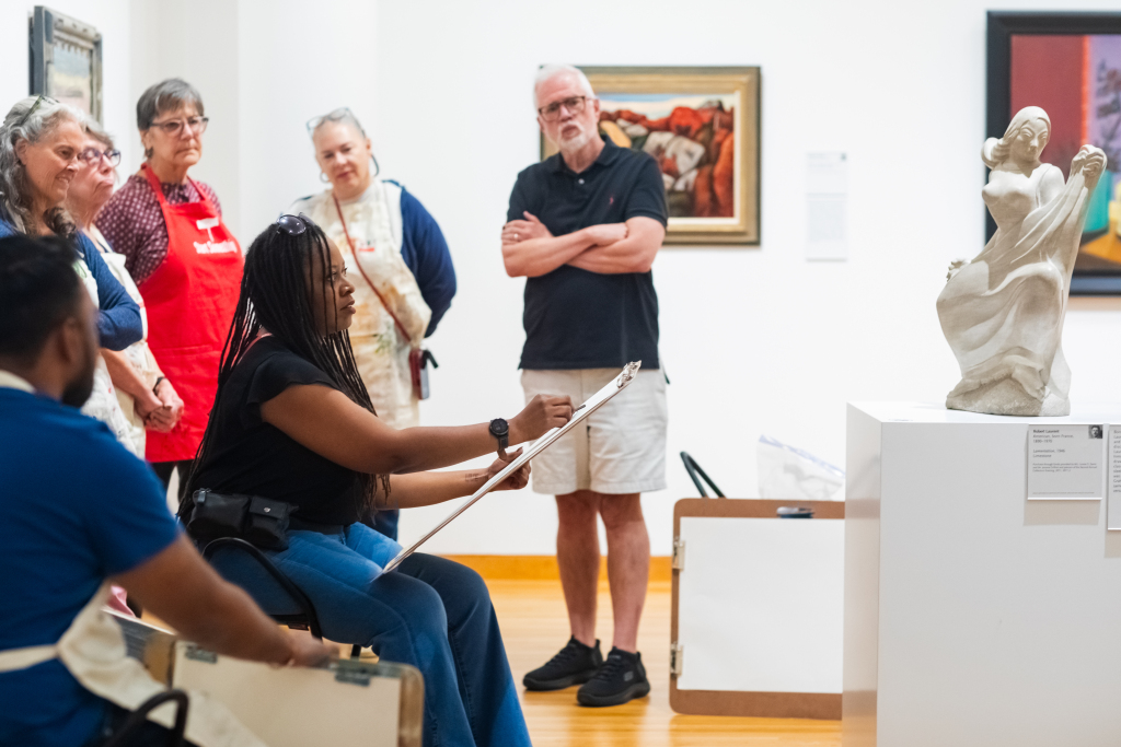 A group of people observes as a woman sketches a sculpture in an art gallery. Several paintings hang on the white walls behind them.