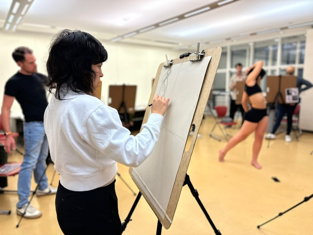 A person sketches on a large easel in an art studio, while a model poses in the background. Other artists and easels are also visible in the bright, spacious room.