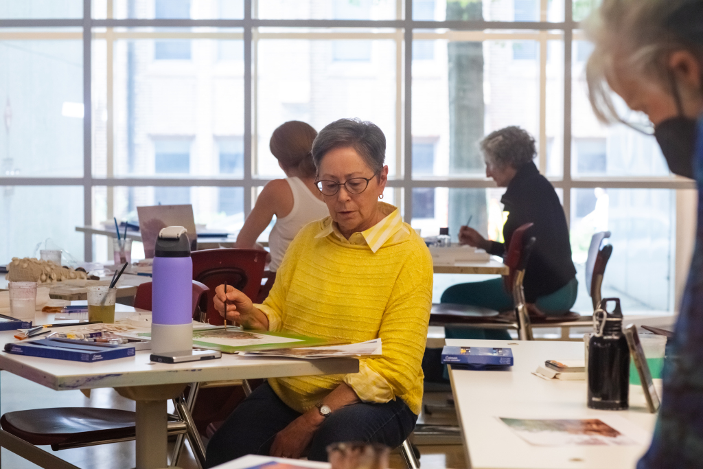 A woman in a yellow sweater sits at a table painting, surrounded by art supplies. Other people work at nearby tables in a bright room with large windows.