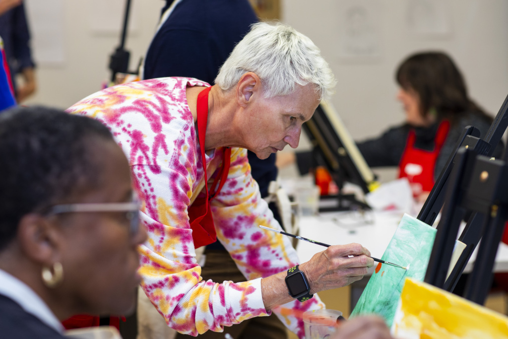 A person with short gray hair paints on a canvas at an art class, wearing a colorful shirt and red apron. Others are painting in the background.