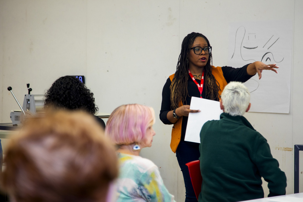 A woman stands and gestures while speaking to a seated group in a classroom. She holds papers and wears glasses and a yellow vest. A drawing is on the wall behind her.