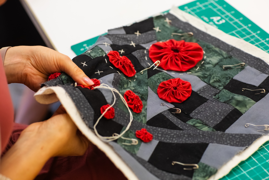 A person holds a fabric patchwork quilt decorated with red fabric flowers, safety pins, and hand-stitched details over a green cutting mat.