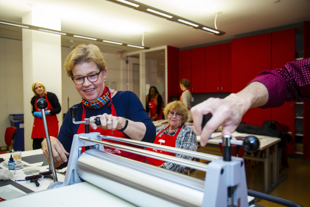 Several adults in a bright room use a printmaking press. One woman smiles while operating the machine. Others watch or assist, and everyone wears red aprons. The atmosphere is friendly and creative.