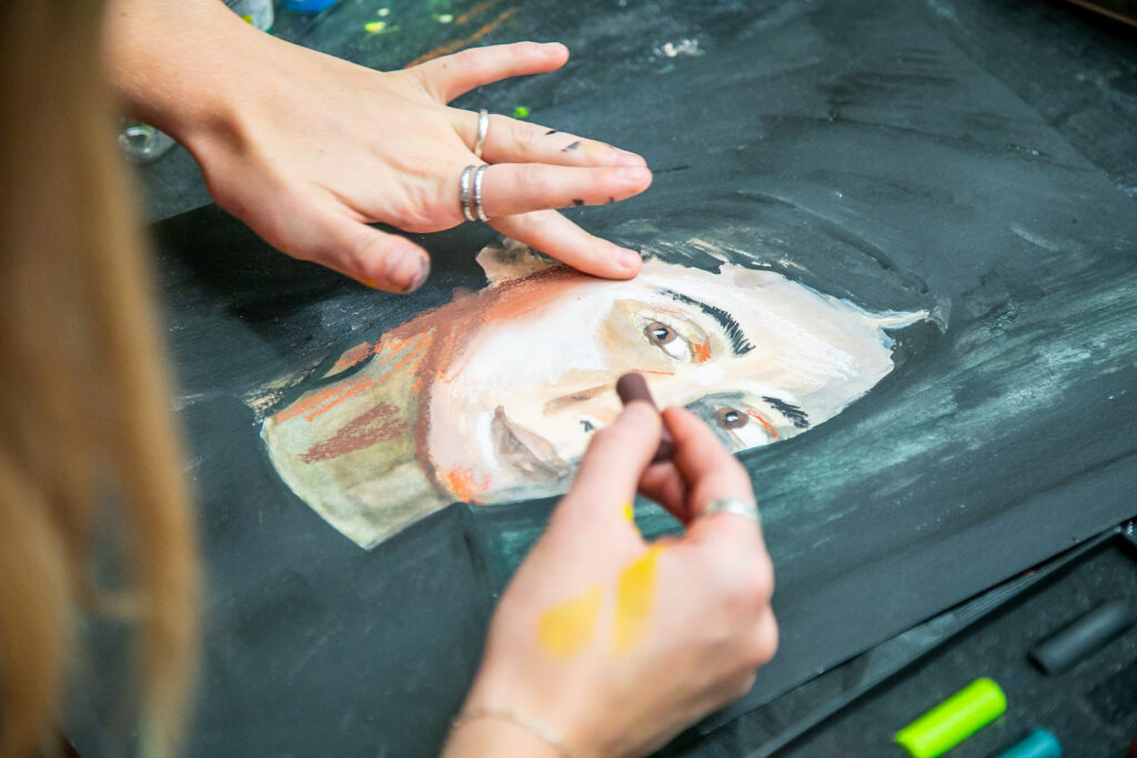 A person draws a portrait of a man's face with pastels on black paper. Their hands, with rings, blend and apply colors, while art supplies are visible nearby.