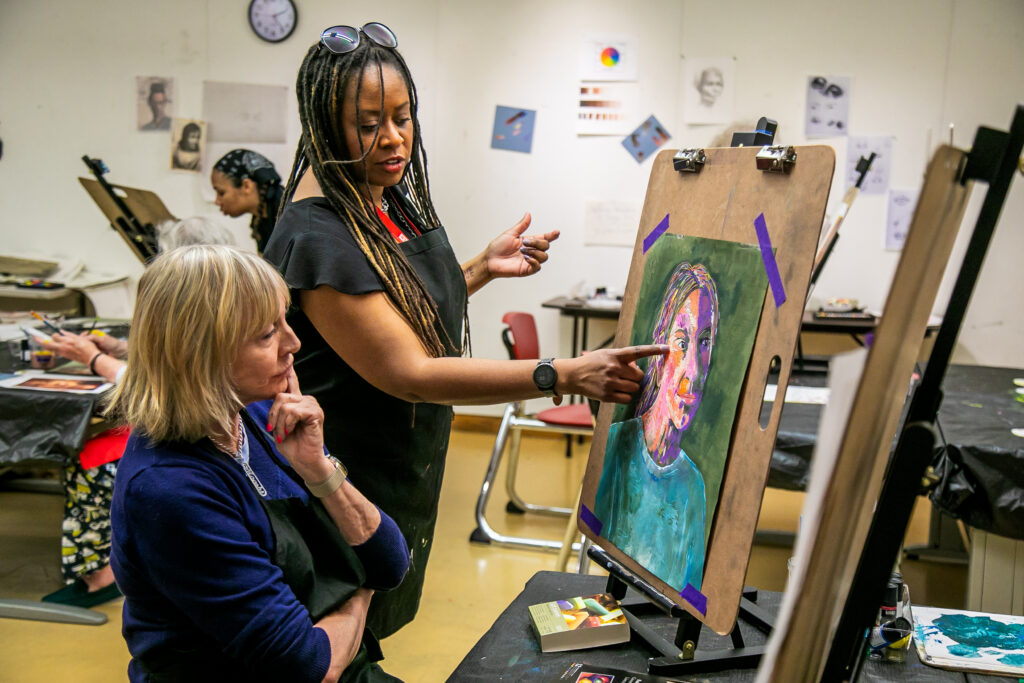 An art teacher points at a student’s colorful portrait painting on an easel in a classroom. Other students work in the background surrounded by sketches and art materials.