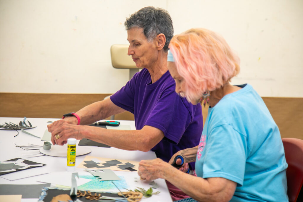 Two older adults sit at a table, creating crafts with paper, glue, and scissors. They focus intently on their projects, surrounded by art supplies, in a bright, casual setting.