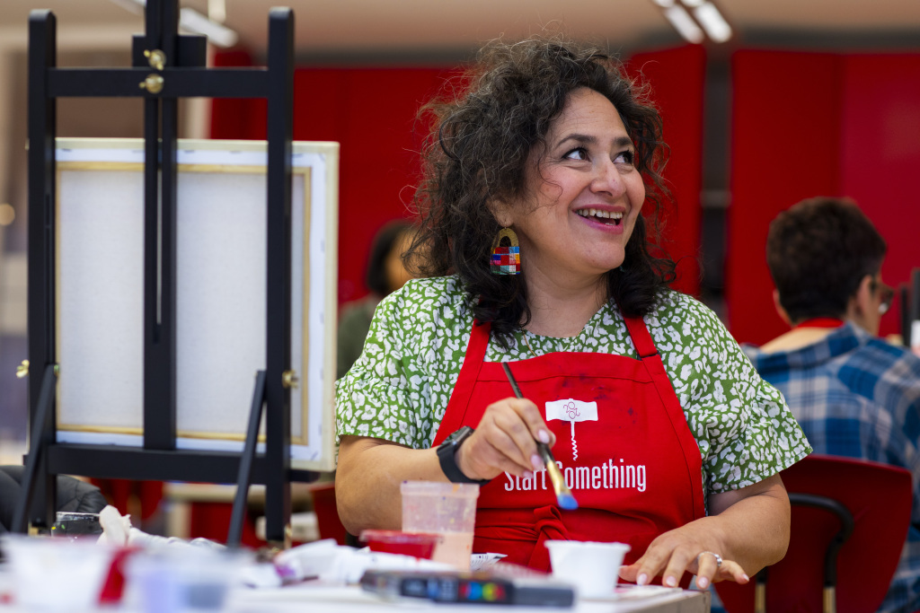 A woman with curly hair wearing a red apron and colorful earrings smiles while painting at a table, surrounded by art supplies. Other people are visible in the background.