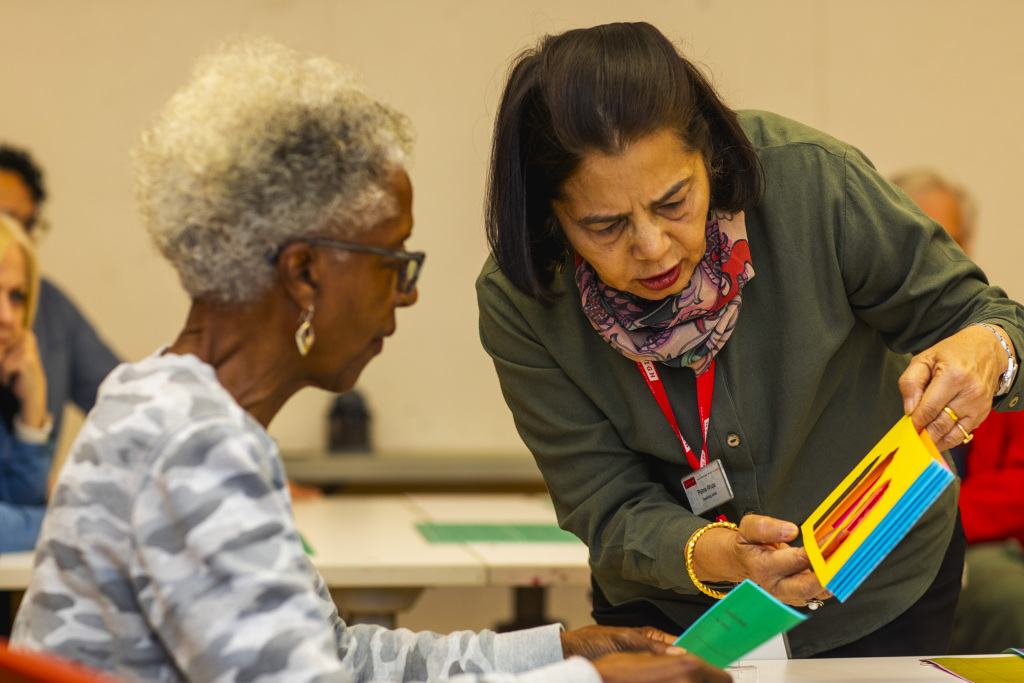 An instructor leans over to help an older woman with colorful cards in a classroom setting. Other participants are seated in the background, focusing on their own tasks.